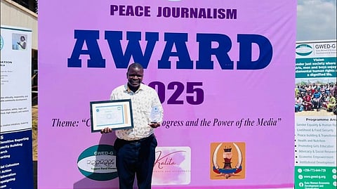 A person smiles while holding a framed certificate in front of a large banner reading "Peace Journalism Award 2025." The scene conveys a sense of achievement and celebration.