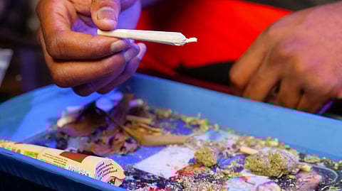 A hand holds a rolled joint above a blue tray filled with cannabis crumbs, grinder, and rolling papers.