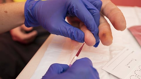 A healthcare professional in blue gloves uses a small tube to collect blood from a person's fingertip. The setting is clinical and focused.