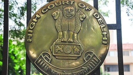 Brass emblem of the Delhi High Court on a gate, featuring the Ashoka Lion capital, with greenery and a building in the background, conveying authority.