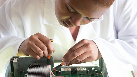 A person in a white lab coat focuses intently on repairing a circuit board, skillfully using tools, conveying precision and technical expertise.