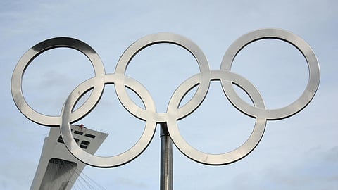 Silver Olympic rings on a pole set against a cloudy sky. A slanted architectural structure appears in the background, adding depth and context.