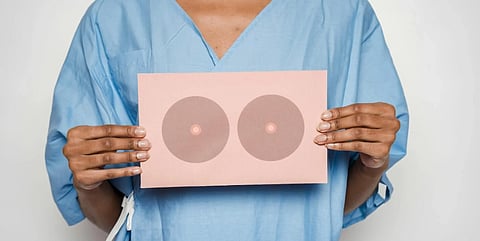 Person in a light blue medical gown holds a pink paper with two circular illustrations symbolizing breasts, conveying breast health awareness.