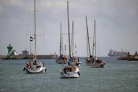 A fleet of sailboats with raised masts heads towards a harbor, under a cloudy sky. A green lighthouse and ships are visible in the background.