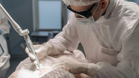 A surgeon, wearing protective gear and a mask, performs a precise procedure using a laser tool in a sterile, focused operating room environment.
