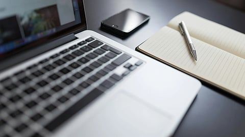 Open laptop on a desk with a smartphone, a lined notebook, and a pen. The scene conveys productivity and organization in a calm workspace.