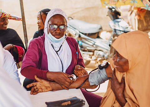 A healthcare worker checks an elderly woman's blood pressure outdoors. The worker is focused, wearing a stethoscope and glasses, while others converse nearby.
