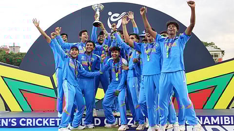 U-19 Indian Cricket team dressed in blue jersey jubilantly celebrating a victory, holding the ICC U-19 World Cup trophy. Smiling faces and raised hands convey joy and triumph.