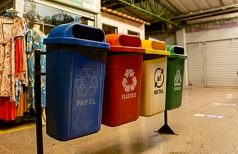 Four recycling bins are lined up indoors: blue for paper, red for plastic, yellow for metal, and green for glass. Clothing is visible on the left.