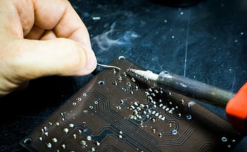 Close-up of a hand soldering components on a circuit board. Smoke rises as the soldering iron applies heat, creating a focused and skillful atmosphere.