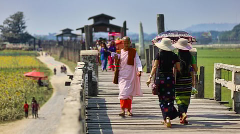 A serene monk in pink and brown robes walks across a wooden bridge, surrounded by green fields. Women with umbrellas stroll nearby under a clear sky.