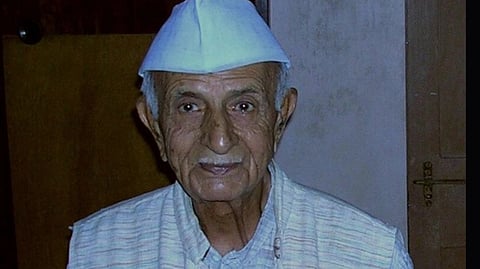 Indian mathematician, theoretical astrophysicist and educationist P.C. Vaidya smiling gently, wearing a white cap and traditional attire, sits against a wooden door backdrop, conveying warmth and wisdom.