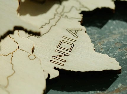 Wooden carving of India's map on a textured surface. The word "India" is engraved prominently, with light and shadow adding depth and focus.