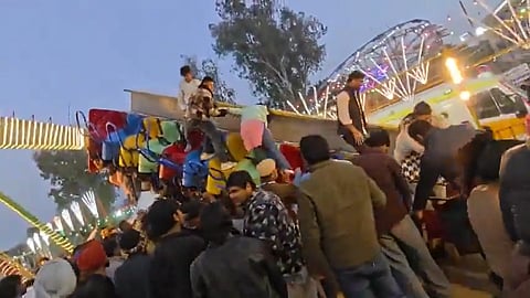 A screenshot from a video showing a crowd around a broken mela swing while people try to climb out.