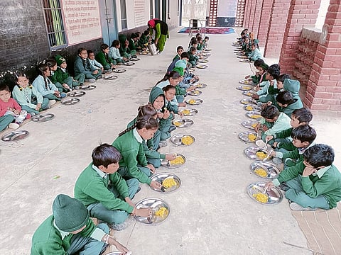 schoolchildren in green uniforms sit in three rows on the ground eating food oout of steel plates.