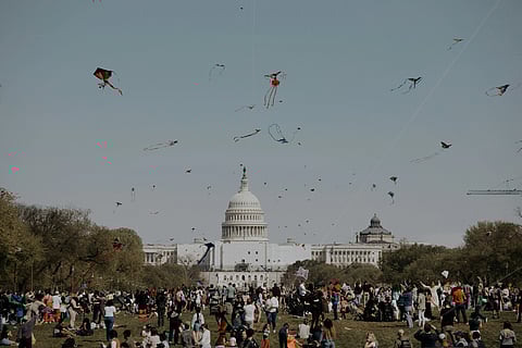 A vibrant outdoor scene with a diverse crowd flying colorful kites on a sunny day near the U.S. Capitol. The atmosphere feels festive and lively.
