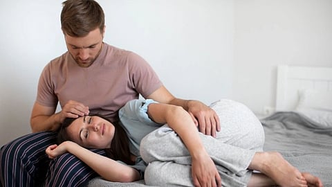 A man in a mauve shirt sits on a bed, gently cradling a woman in gray pajamas resting her head in his lap. The scene conveys comfort and tenderness.