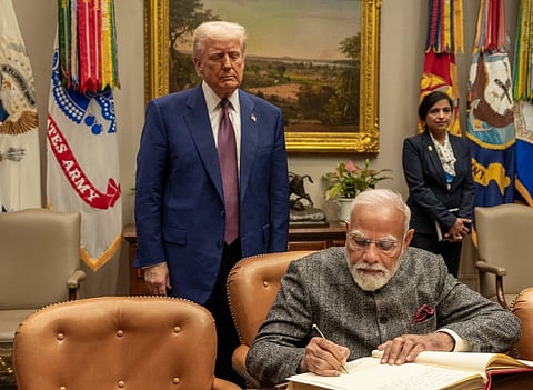 Image of President Donald Trump standing behind PM Modi while he is signing something on the table. A woman stands in the background. Flags and a landscape painting decorate the room.