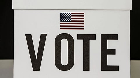 A hand places a ballot into a white box labeled "VOTE," featuring a small American flag. The image conveys a sense of civic duty and participation.