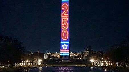The Washington Monument illuminated at night with red and blue lights displaying "250" and stars, reflecting in a calm pool. Celebratory and patriotic mood.
