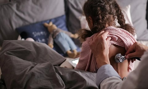A child sitting on a bed with a doctor listening to her back with a stethoscope. The room is cozy, with pillows and a stuffed toy nearby.