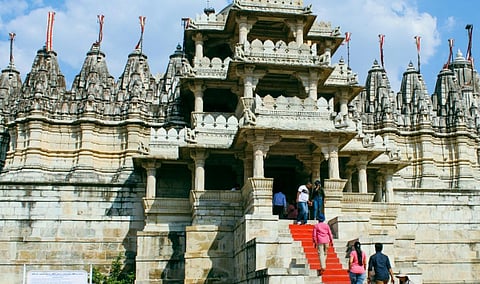Image of ranakpur jain temple with detailed stone carvings, featuring tall spires and red-carpeted steps, under a blue sky. Visitors explore the entrance, evoking awe.