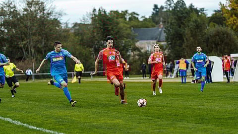 Two soccer teams compete on a grassy field. A player in red actively dribbles the ball while players in blue and red give chase. The scene is dynamic and competitive, with a focused atmosphere.