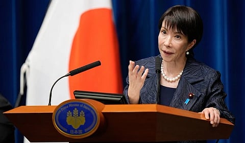 Sunae Takaichi in formal attire speaks at a podium with the Japanese flag in the background. She appears focused and composed, conveying authority and confidence.