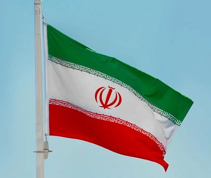The image shows Iran's flag waving against a clear blue sky. The flag has horizontal green, white, and red stripes with an emblem in the center.