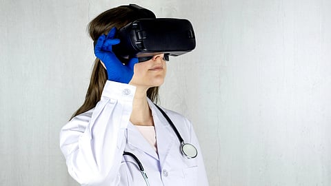 A doctor wearing a VR headset and blue gloves, dressed in a white coat with a stethoscope, stands against a plain white background.