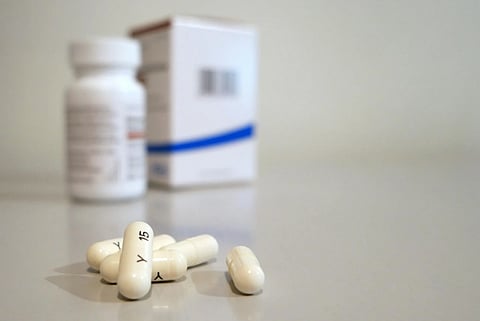 White capsules with markings are in focus on a smooth surface, with a blurred pill bottle and box in the background.