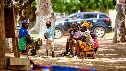 A group of women and children sit on stools in a sun-dappled park under large trees. A black SUV and colorful clothing create a vibrant, relaxed atmosphere.