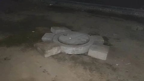 Round manhole cover secured with four large bricks on a dirt ground. The scene is dimly lit, evoking a sense of nighttime scenery and solitude.