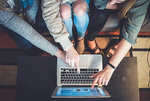 Three people sit on a sofa, pointing at a laptop screen with engaged expressions, suggesting teamwork or collaboration. Casual attire reflects a relaxed setting.