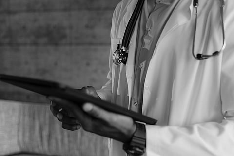 Black and white image of a doctor in a white coat, holding a tablet. A stethoscope hangs around their neck. 