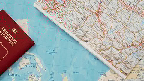 A red passport lies on a colorful world map beside a folded road map. The scene suggests travel, exploration, and international journey plans.
