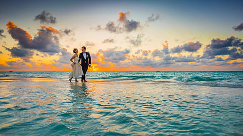 A couple walks on a beach during a vibrant sunset, the sky painted with orange and purple hues. They wear wedding attire, evoking romance and joy.