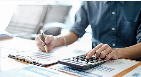 A person in a blue shirt uses a calculator on a desk filled with charts and graphs. They hold a pen, conveying a focus on financial analysis.