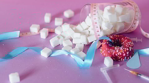 Scattered sugar cubes, measuring tape, blue ribbon, syringe, and a sprinkled donut on a pink background evoke themes of sweetness and health.