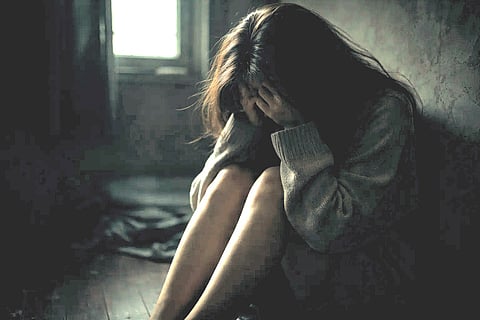 A woman in a dimly lit room sits on the floor, hugging her knees and covering her face, conveying feelings of sadness and isolation.