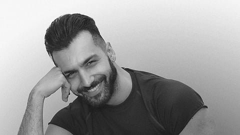 Black and white image of a smiling man with a beard, resting his head on his hand. He exudes a relaxed and confident vibe, wearing a casual t-shirt.