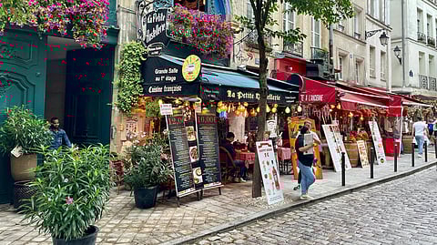 A lively Parisian street with charming cafés, colorful signs, hanging flowers, and cobblestone paths. People dine outside, creating a vibrant, cozy atmosphere.