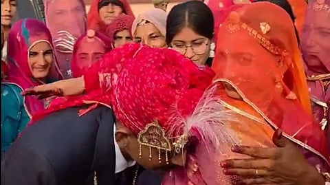 A groom in a colorful turban receives a blessing from a woman in traditional attire, surrounded by smiling family members in vibrant saris. 
