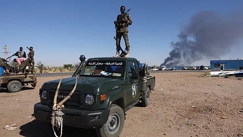 A soldier stands on a green military truck with a mounted gun, against a clear sky. Smoke billows in the distance, conveying a tense and alert atmosphere.
