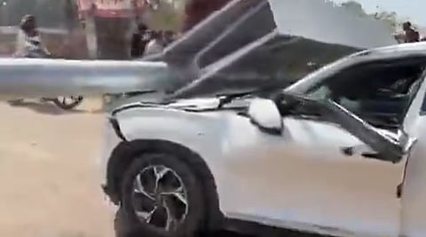 A white car with severe damage is impaled by a large metal beam on a busy street. The scene appears chaotic, with onlookers in the background.