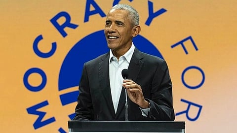 Barack Obama in a suit speaks at a podium with a microphone, smiling. The backdrop reads "Democracy Forum" with an abstract logo. The setting is formal and engaging.