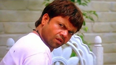 Rajpal Yadav in a white shirt sits outdoors with a puzzled and slightly amused expression. Behind him, there is a decorative white railing and greenery.