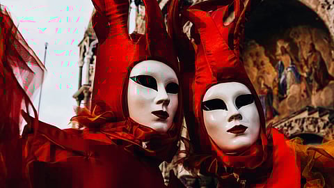 Two figures in striking red costumes and masks with elongated headpieces stand against an ornate background, evoking a mysterious, theatrical atmosphere.