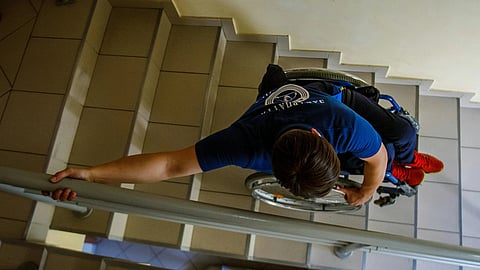A person in a wheelchair navigates down a tiled staircase, with one hand on the railing. The focus is on the action and determination.