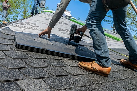 Worker in jeans and boots installs roof shingles with a nail gun on a sunny day. The scene conveys focus and precision in construction work.